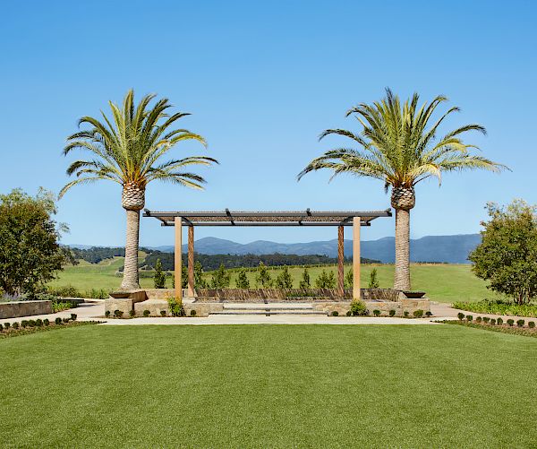 A well-manicured lawn with a pergola flanked by palm trees in a scenic outdoor area, surrounded by greenery and a mountainous horizon.