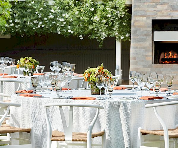An outdoor dining setup features white tablecloths, floral centerpieces, wine glasses, and neatly arranged place settings; a fireplace is visible in the background.