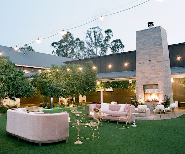 An outdoor seating area features pink couches, a tall stone fireplace, and string lights, set in a garden with trees and greenery surrounding it.
