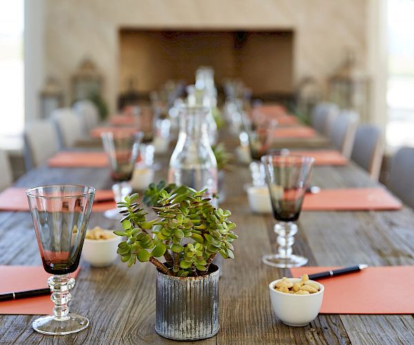 The image shows a long wooden table set for a meeting, with glasses, notebooks, pens, a potted plant, snacks, and a water carafe.