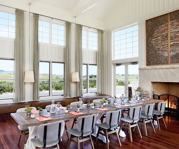 A spacious dining room with a long table, several chairs, tall windows, a fireplace, and a scenic view of the outdoors.