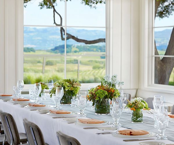 This image shows a long dining table set with plates, glasses, and floral centerpieces, in a room with large windows overlooking a scenic landscape.