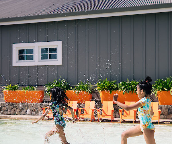 Two kids play in a shallow pool surrounded by orange chairs and planters, near a gray building with a window.