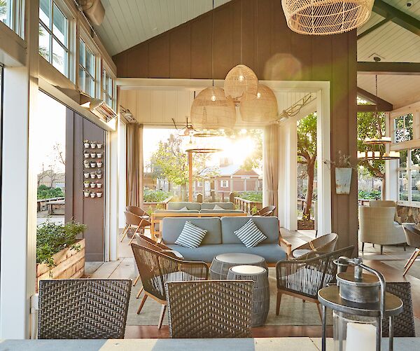 A sunlit patio with cozy seating, wicker chairs, hanging lights, and sun streaming through large windows, offering a warm, inviting ambiance.