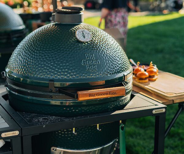 A green ceramic grill on a stand with a thermometer is in a garden setting. A cutting board with food is nearby.