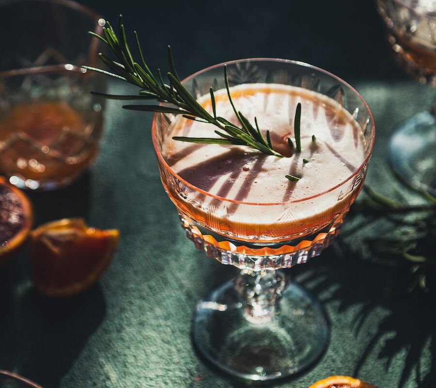 A cocktail garnished with rosemary in a glass, surrounded by citrus slices and additional garnishes on a dark surface.