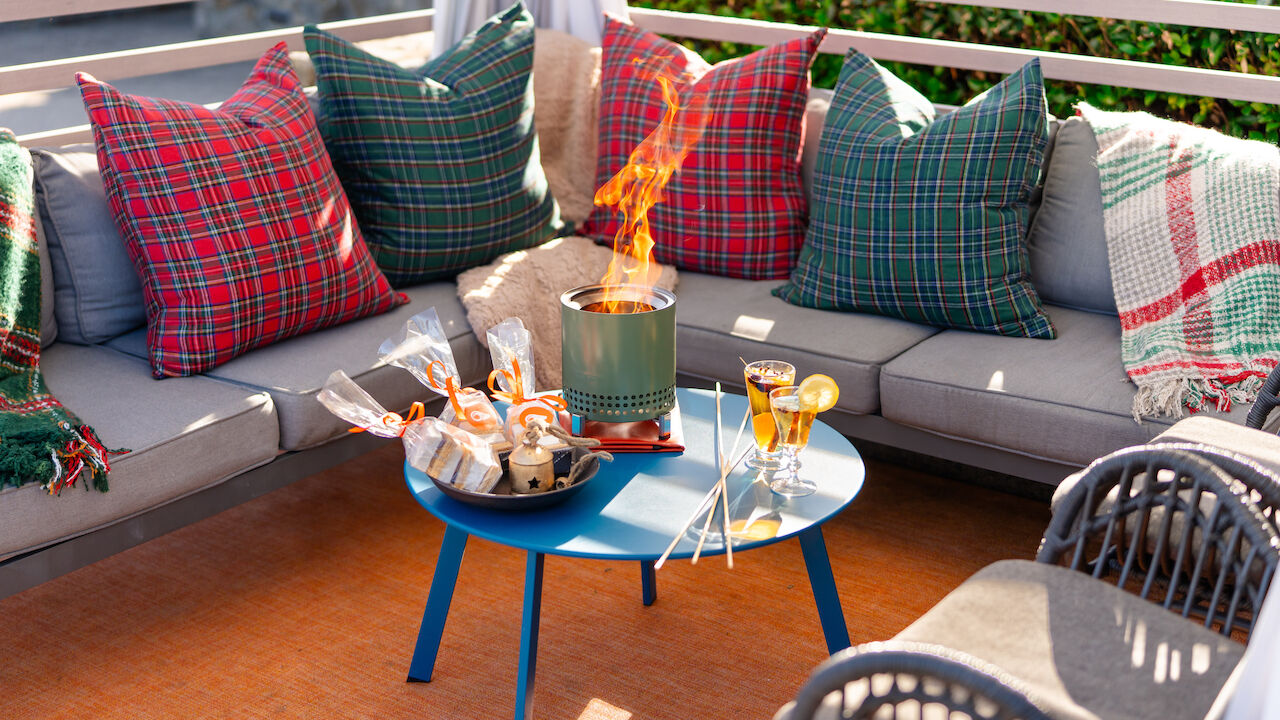 A cozy outdoor seating area with plaid pillows, a small fire pit, drinks, and snacks on a blue table.