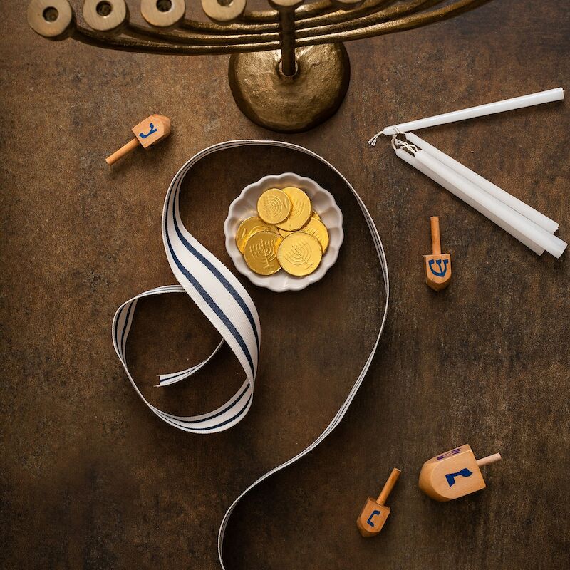 A menorah, dreidels, wrapped candles, a ribbon, and a bowl of coins are arranged artfully on a dark surface.
