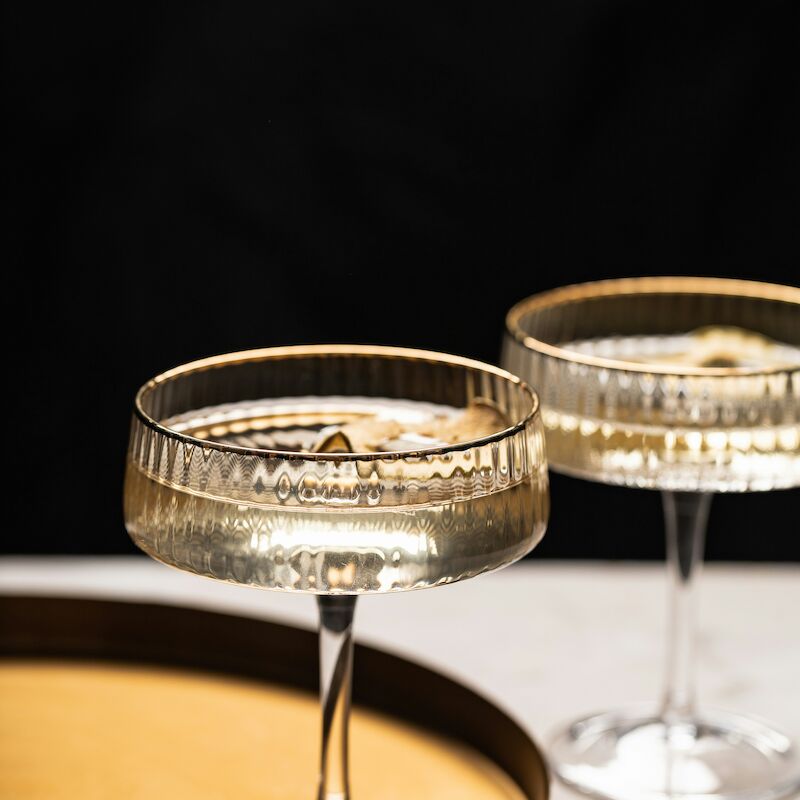 Two elegant champagne coupes filled with bubbly liquid are set against a dark background, accompanied by a golden tray.