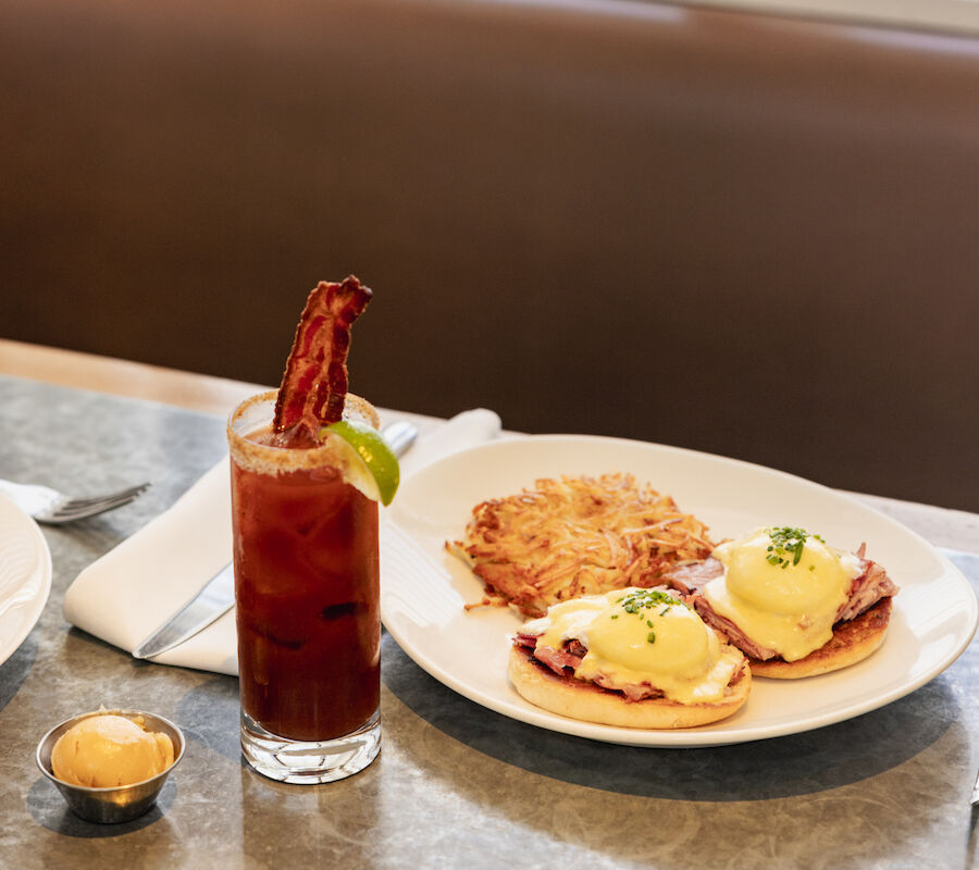 A breakfast setup with eggs Benedict, hash browns, and a drink garnished with bacon and a lime slice, on a table.
