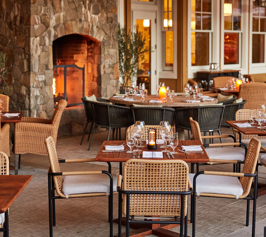 The image shows a cozy outdoor restaurant setting with a stone fireplace, wicker chairs, and tables set for dining with wine glasses and candles.