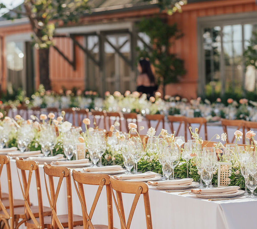 A long, elegantly set outdoor dining table with wooden chairs, floral centerpieces, and glassware under trees in a garden setting.