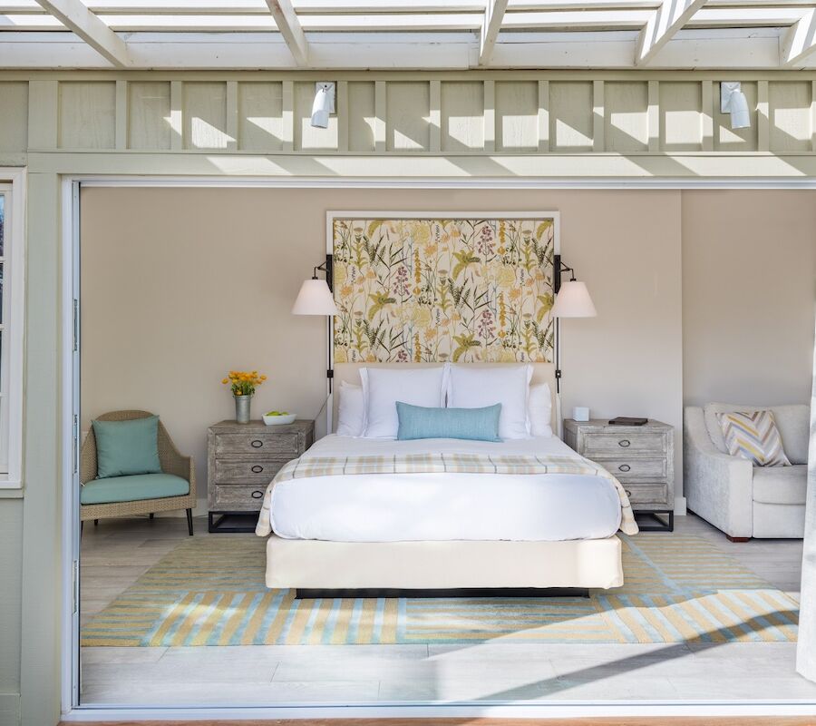 A cozy bedroom with a large bed, decorative pillows, side tables, and a patterned artwork, in a room with natural light.