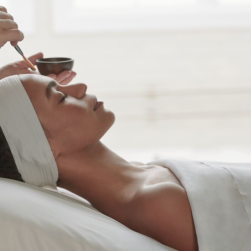A person is lying down receiving a facial treatment, with a brush applying a mask, and a headband keeping hair away.