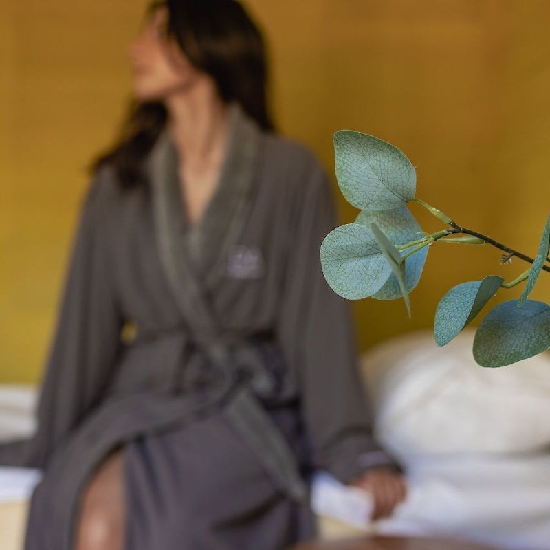 A person in a robe sits indoors with eucalyptus leaves in focus, creating a relaxed atmosphere.