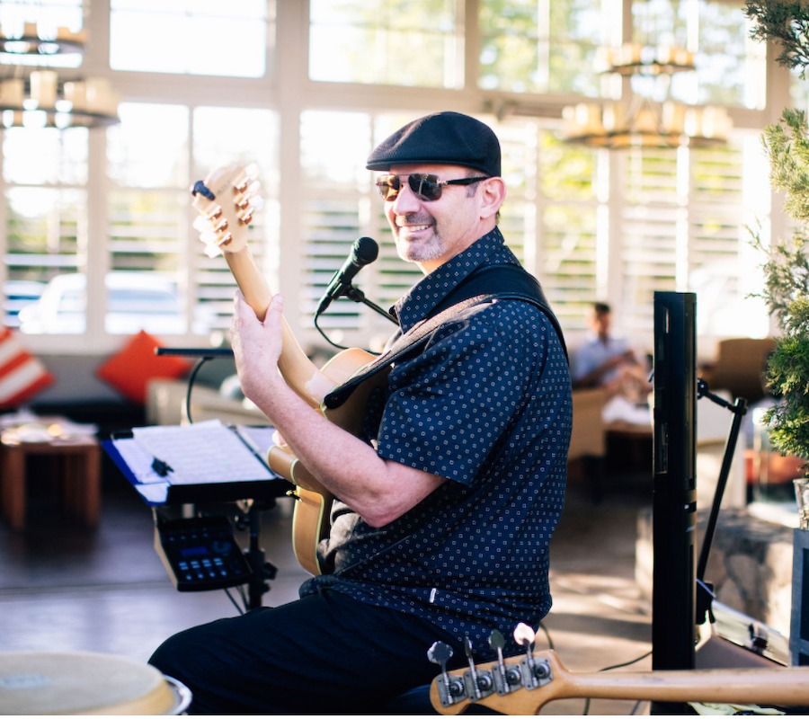A musician, wearing a cap and sunglasses, smiles while playing an electric guitar with a microphone in front of him at what seems to be an outdoor venue.