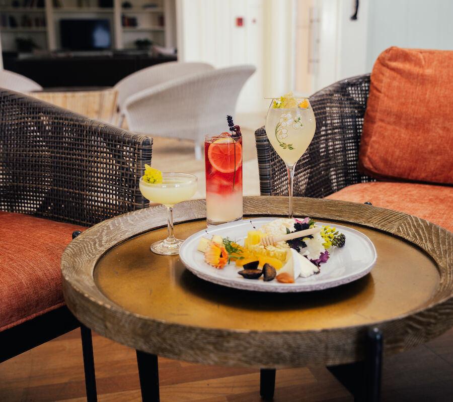 A table with a cheese platter and three cocktails between orange chairs in a cozy setting.