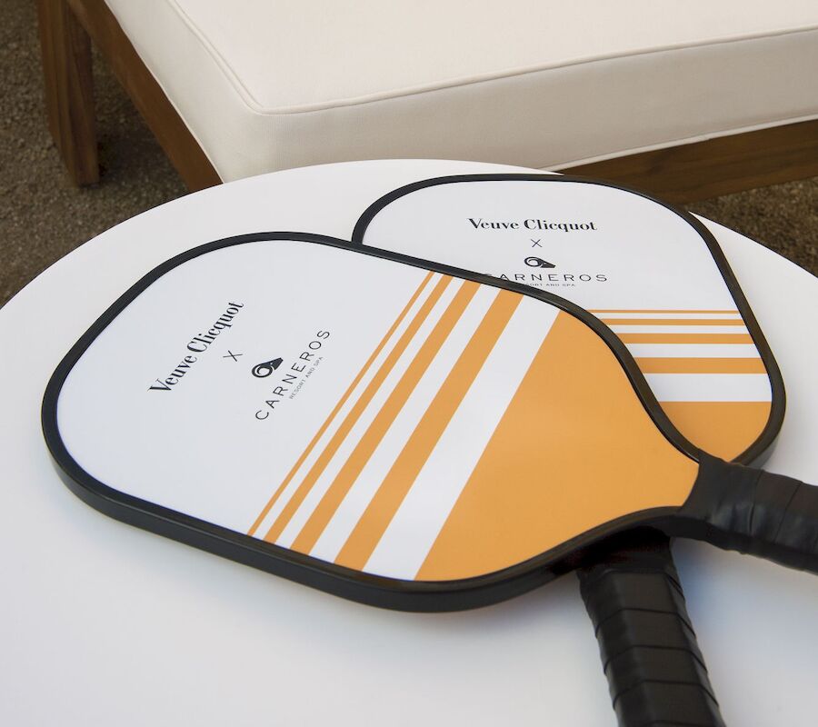 Two pickleball paddles with a white and orange design on a round white table, with a cushioned bench in the background.