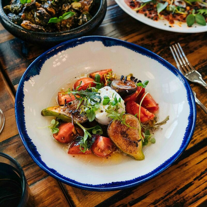 A delicious dish with figs, burrata, and greens on a table set with cutlery and wine glasses, creating a cozy dining ambiance.