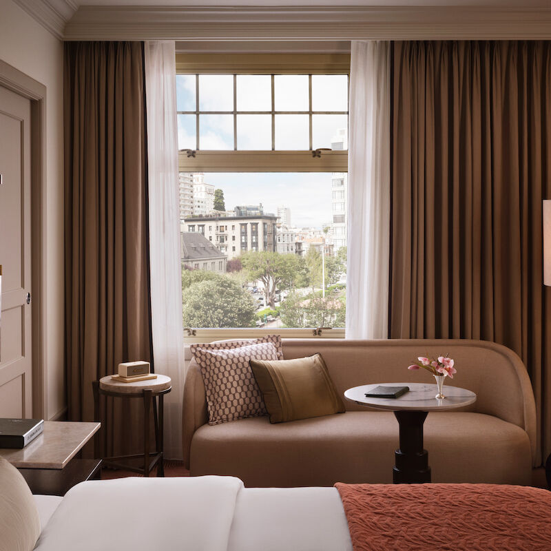 A cozy hotel room with a bed, sofa, and table by a window showcasing an urban view, complete with warm brown curtains.