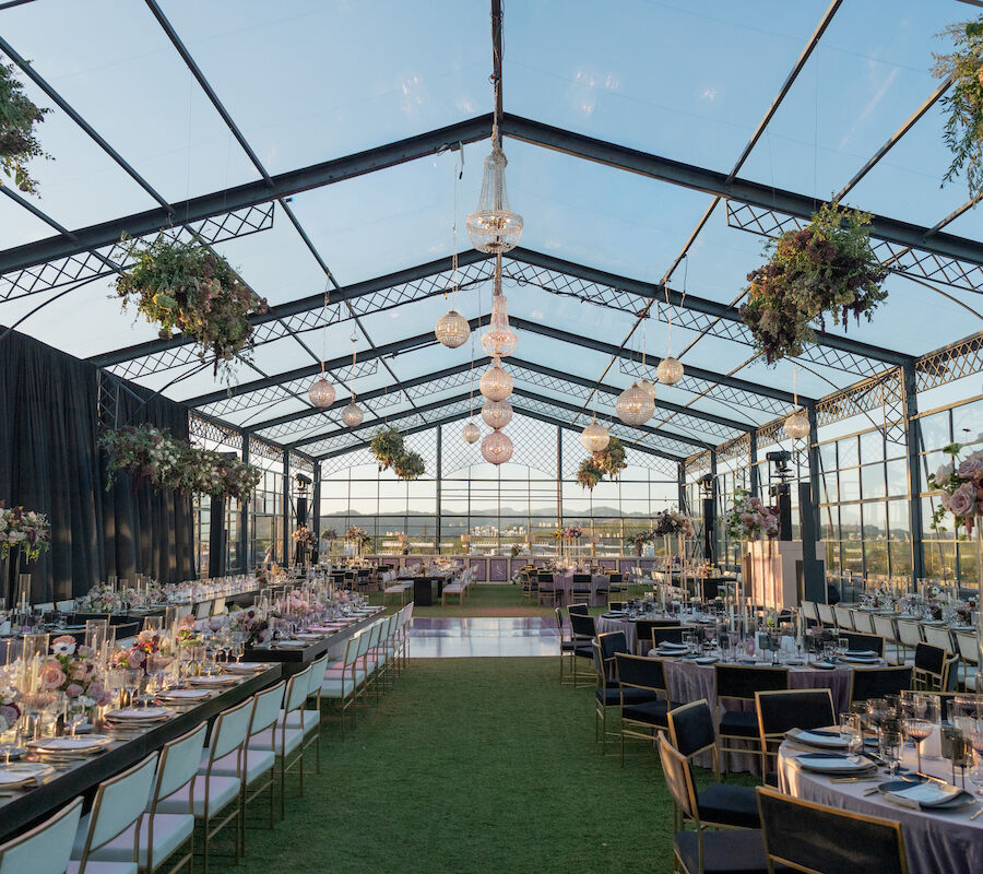 The image shows a beautifully decorated event space with tables, chairs, hanging lights, and greenery under a large glass canopy.