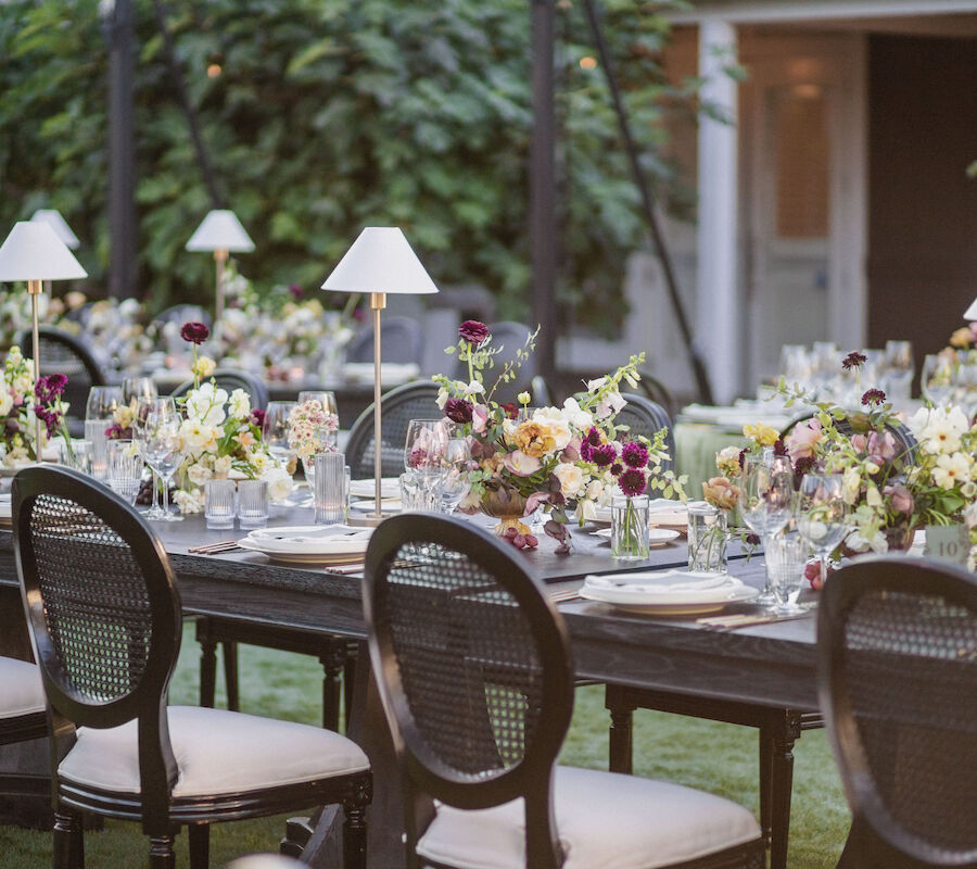 An elegantly set outdoor dining table with floral arrangements, table lamps, and chairs.