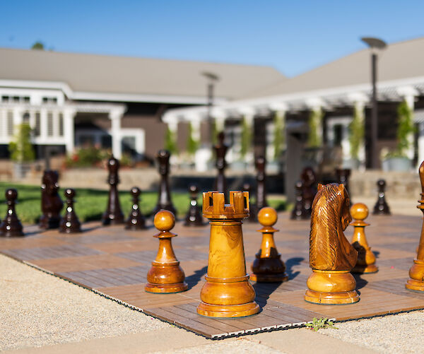 The image shows a large outdoor chess set with wooden pieces on a board, set in a sunny courtyard area.