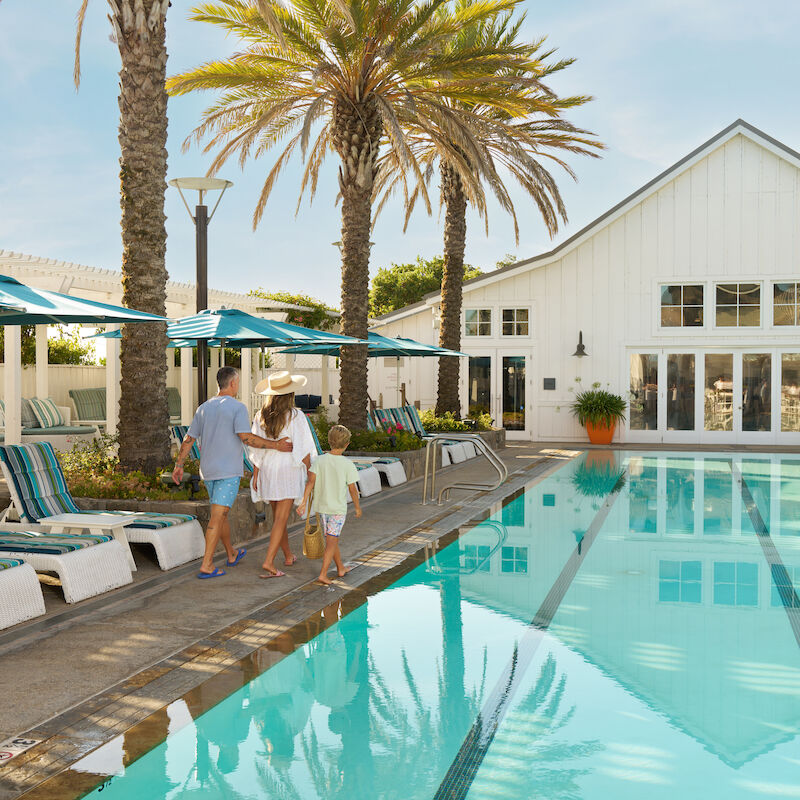 A family walking beside a pool with palm trees and a white building in the background.