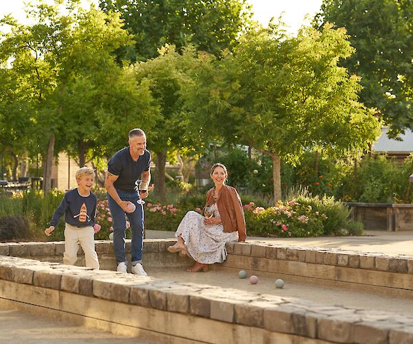 A group enjoying a game of bocce ball outdoors on a sunny day near trees and flowerbeds.