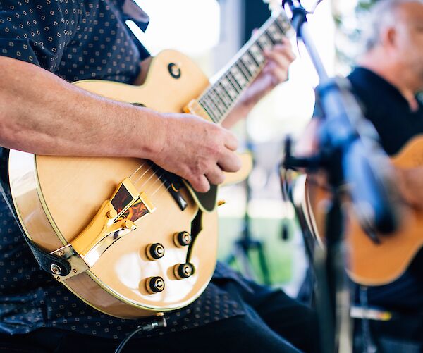 Two individuals are playing guitars, one in the foreground and another in the background, with a focus on their instruments.