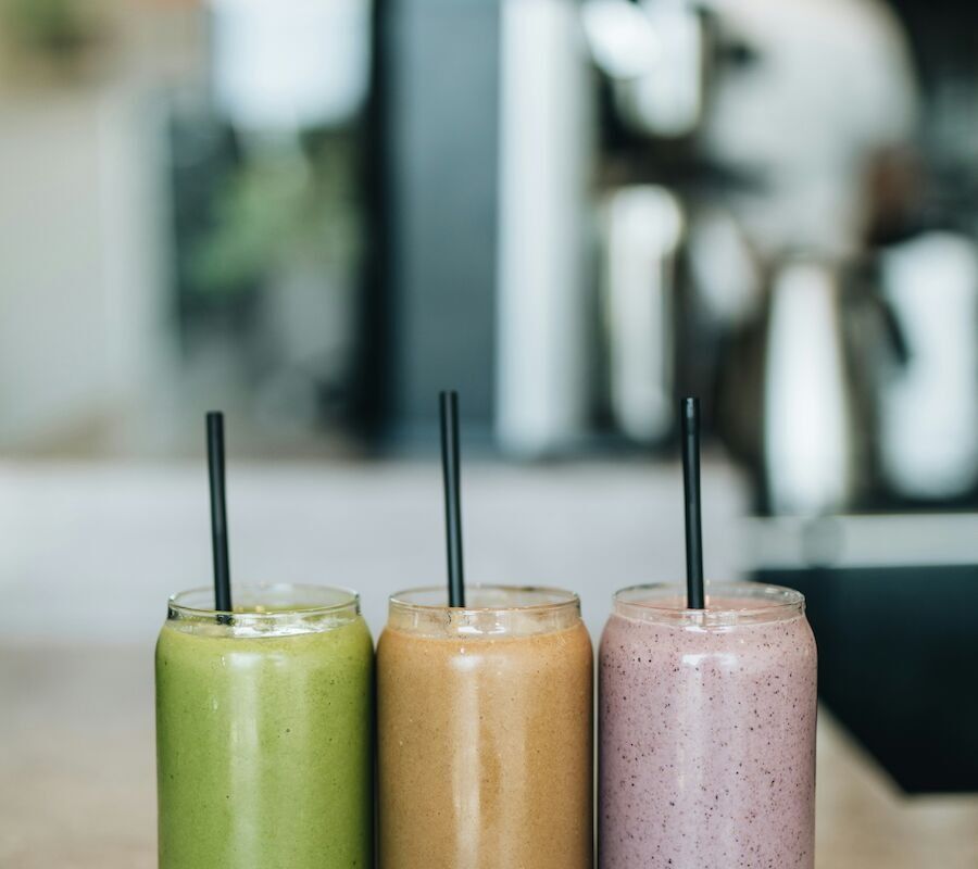 Three smoothies in glasses with black straws in various colors—green, orange, and pink—are placed on a countertop.