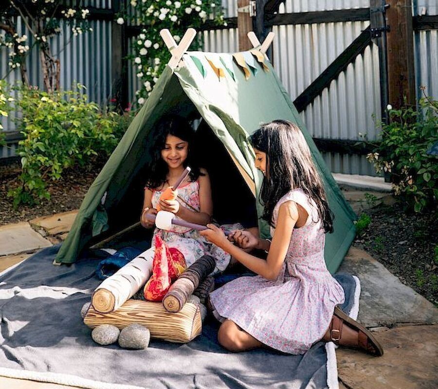 Two kids are sitting near a small green tent in a backyard, playing with sticks and toys on a mat.