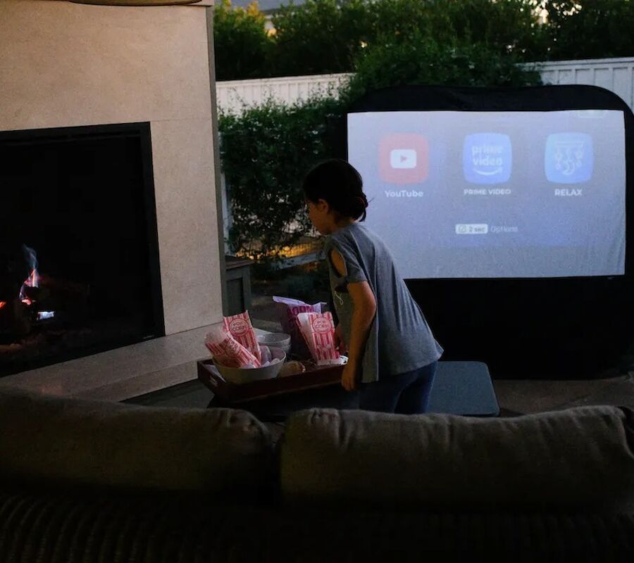 An outdoor setup with a person carrying popcorn near a fireplace and a screen displaying video app icons.