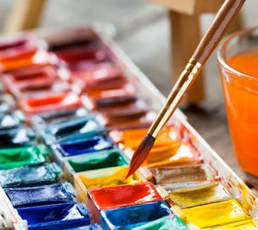 A watercolor paint set with various colors is shown alongside a brush and a glass of orange liquid.