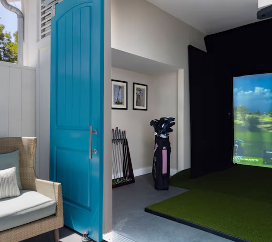 The image shows a room with a golf simulator on the right, a set of golf clubs, and a cozy chair by a blue door.