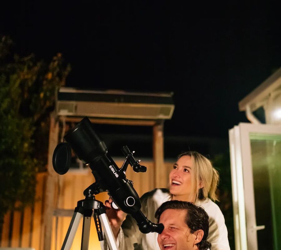 A couple in robes is stargazing, using a telescope at night. They appear happy and engaged with the activity.