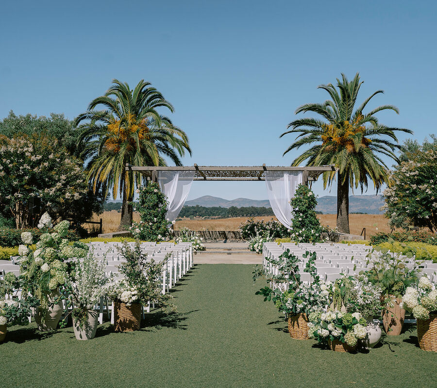 An outdoor wedding setup with white chairs, floral arrangements, and a canopy under palm trees.