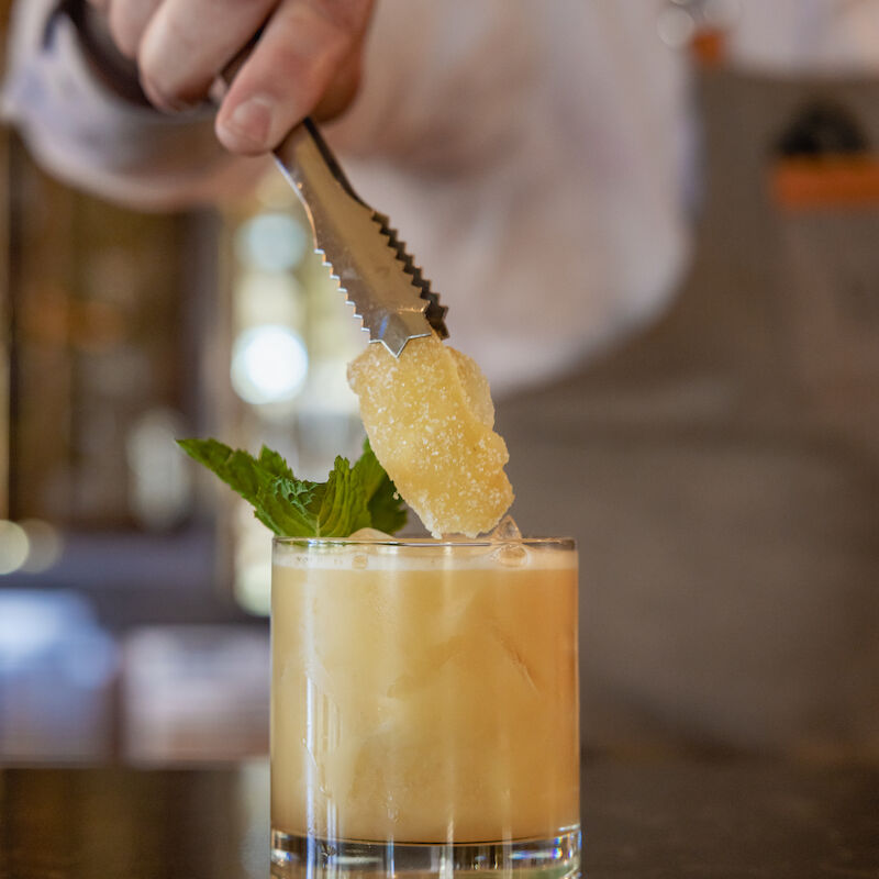 A bartender is adding a garnish with tongs to a cocktail in a glass, which is decorated with a leaf, on a bar counter.