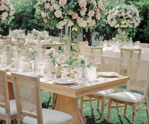 An elegantly set outdoor wedding table with ornate chairs and large floral centerpieces featuring pink and white flowers on a wooden surface.