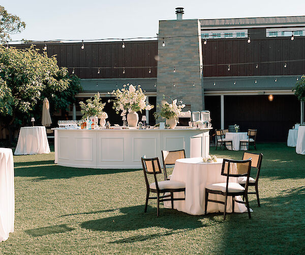 An outdoor event setup with round tables, chairs, a bar with floral arrangements, and string lights overhead on a sunny day.