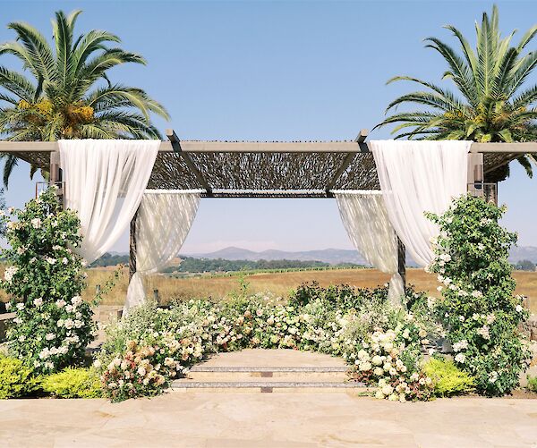 An outdoor wedding arch decorated with white fabric, flowers, and greenery, set between two palm trees under a clear blue sky.