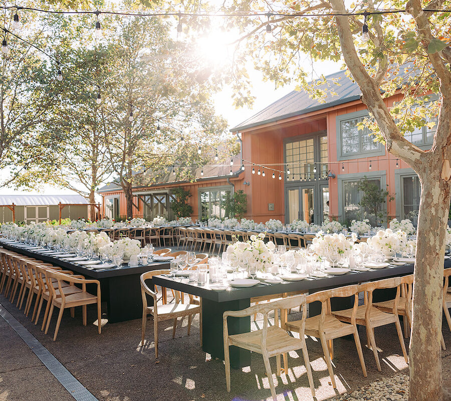 An outdoor event setup features long tables with elegant place settings and floral arrangements, set against a picturesque building backdrop.