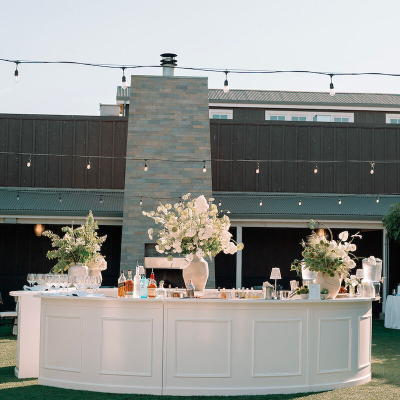 An outdoor event setup with a curved bar, adorned with floral centerpieces, bottles, and glasses, set against a building backdrop.