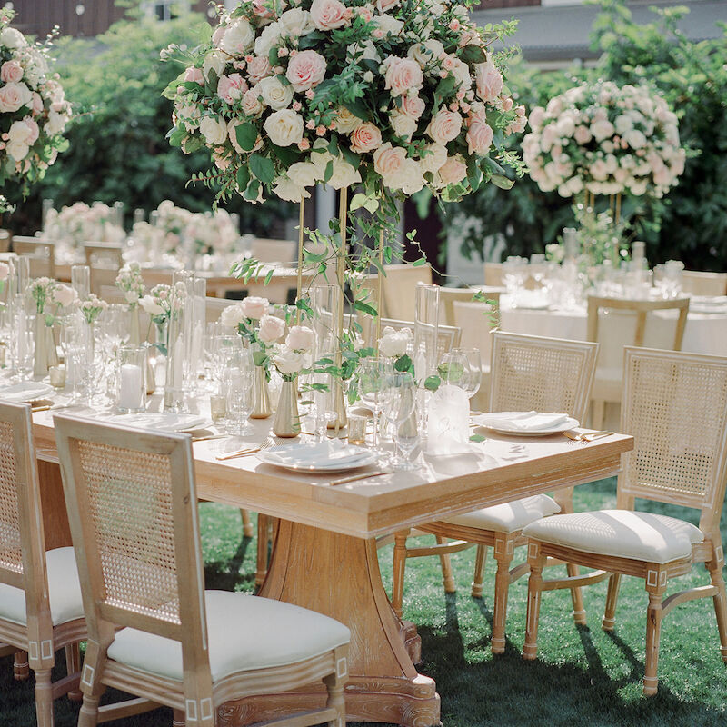 Elegant outdoor event setup with wooden tables, beige chairs, and tall floral centerpieces.