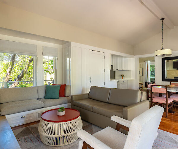 The image shows a cozy living room with sofas, a round red coffee table, and a dining area in the background, featuring large windows.