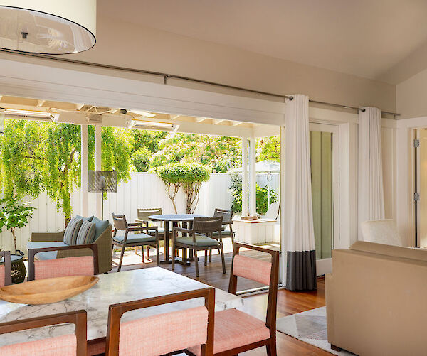 A bright living area opens to a patio with plants and outdoor seating, featuring a dining table inside and another outside.