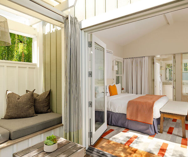 A cozy room with a bed, bench, and colorful rug, next to an open seating area with cushions and a small table holding a plant.