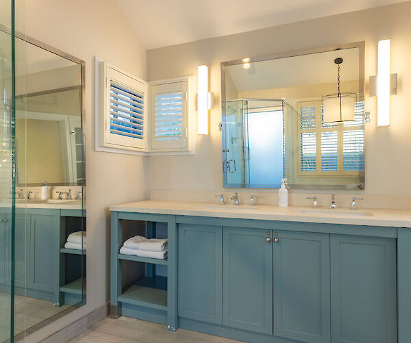 A modern bathroom with a large mirror, blue vanity, two sinks, and shelves with towels. A glass shower enclosure is also visible.