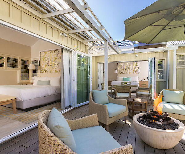 Outdoor seating area with wicker chairs, fire pit, and umbrella; adjacent to two open rooms with beds and seating.