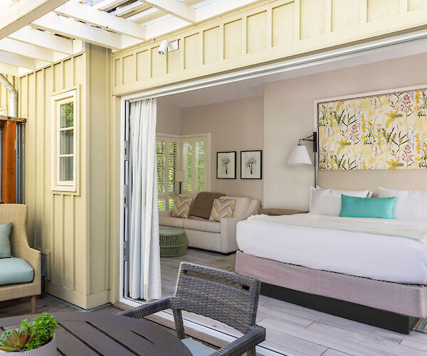 A cozy indoor-outdoor space with a bed, sofa, and chairs under a pergola, featuring soft tones and bright decorations.